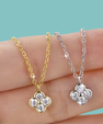 Two flower-shaped pendant necklaces with clear gemstones, one in yellow gold and one in silver, displayed on a person's fingers against a solid teal background.