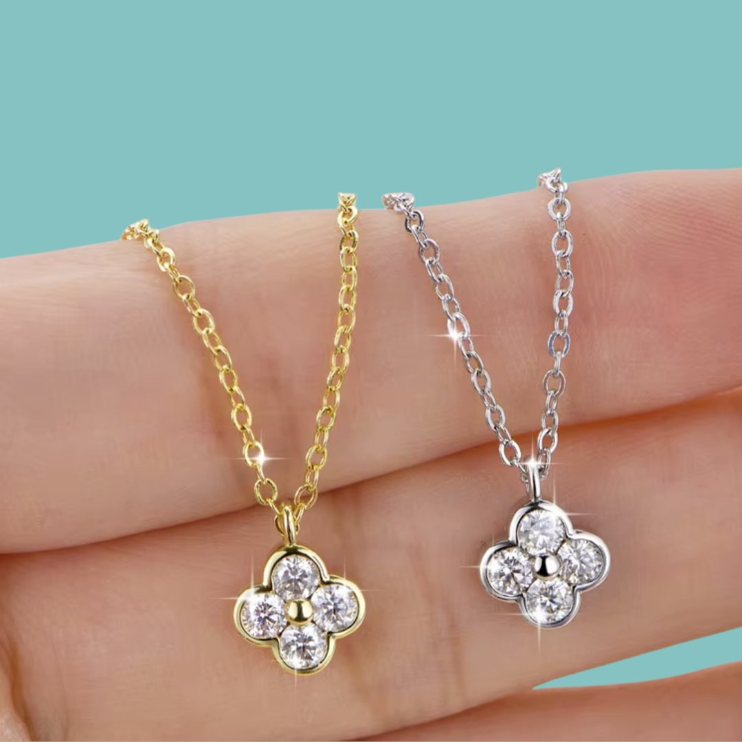 Two flower-shaped pendant necklaces with clear gemstones, one in yellow gold and one in silver, displayed on a person's fingers against a solid teal background.