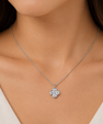A close-up of a woman's neck and chest wearing a delicate silver chain necklace with a small four-petal flower pendant set with clear stones.