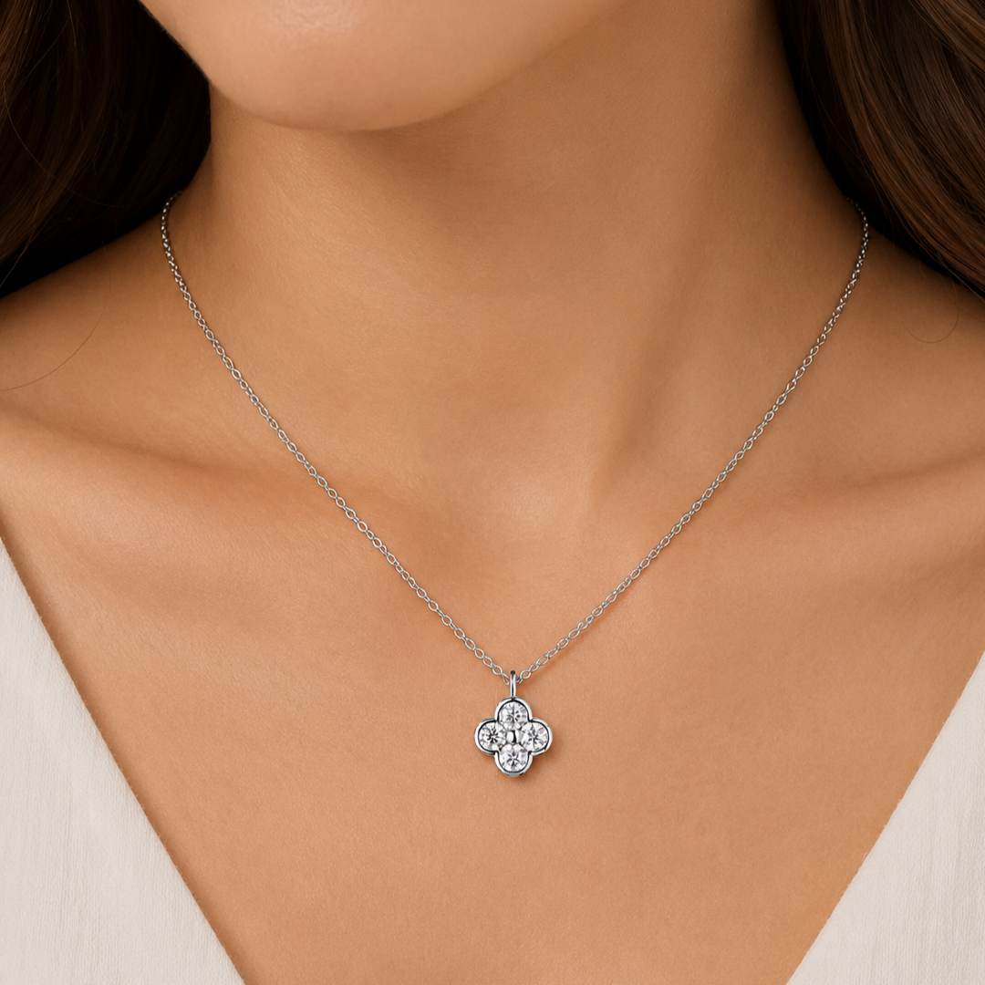 A close-up of a woman's neck and chest wearing a delicate silver chain necklace with a small four-petal flower pendant set with clear stones.