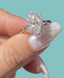 A silver halo ring with a marquise-cut center stone and surrounding round stones, shown being held between fingers against a solid light aqua background.