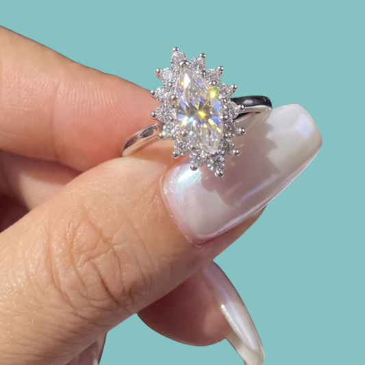 A silver halo ring with a marquise-cut center stone and surrounding round stones, shown being held between fingers against a solid light aqua background.