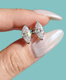 A close-up of a person's hand with long, manicured nails holding a pair of silver stud earrings with marquise-cut clear gemstones against a solid teal background.
