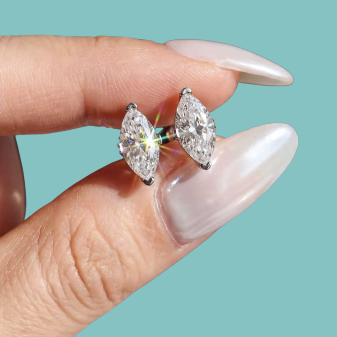A close-up of a person's hand with long, manicured nails holding a pair of silver stud earrings with marquise-cut clear gemstones against a solid teal background.