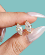 A close-up of a hand with long, glossy pale pink manicured nails holding a pair of gold stud earrings with marquise-cut clear gemstones against a solid light teal background.