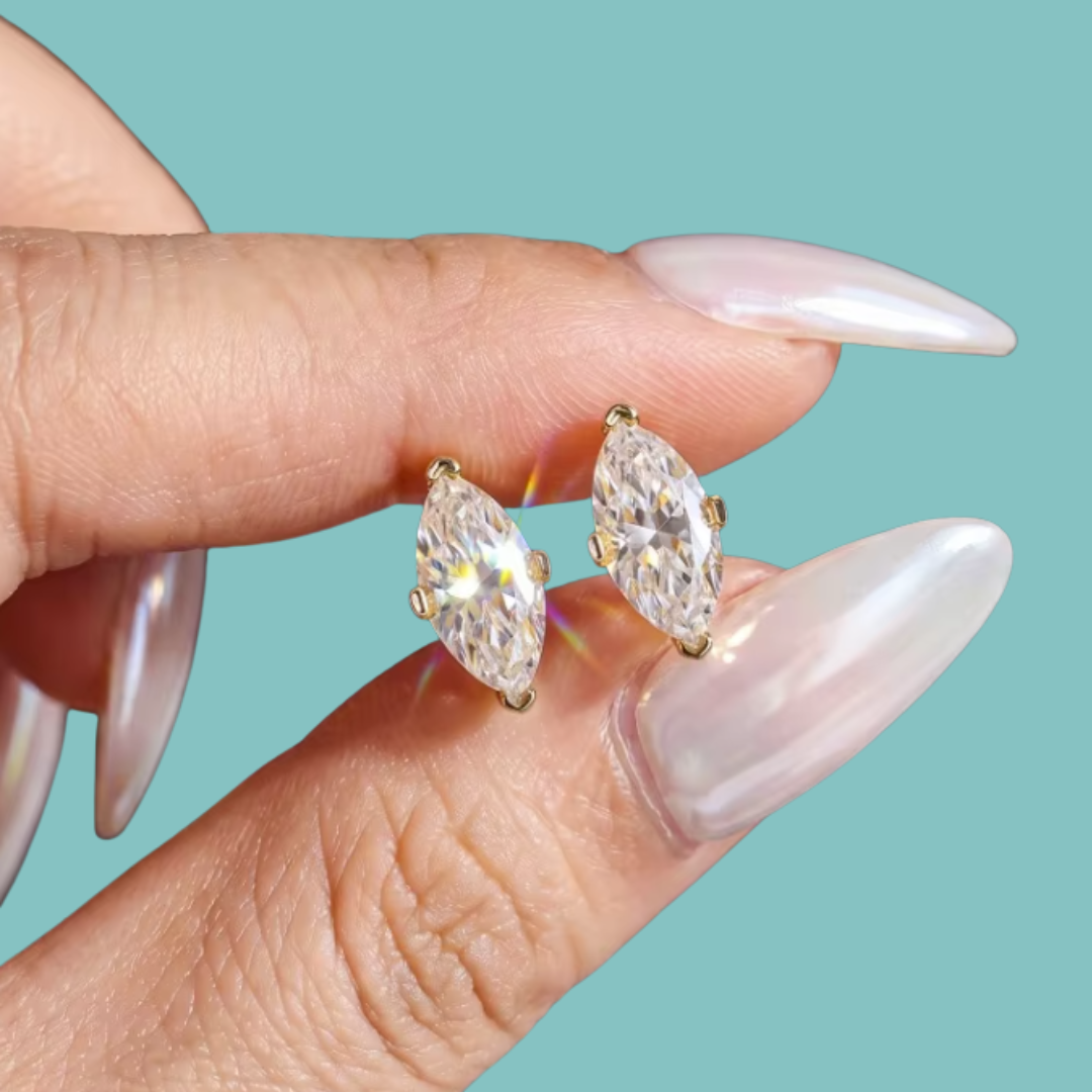 A close-up of a hand with long, glossy pale pink manicured nails holding a pair of gold stud earrings with marquise-cut clear gemstones against a solid light teal background.