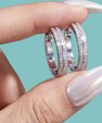 A close-up of a hand with long, glossy white manicured nails holding two silver hoop rings set with small clear stones against a solid light teal background.