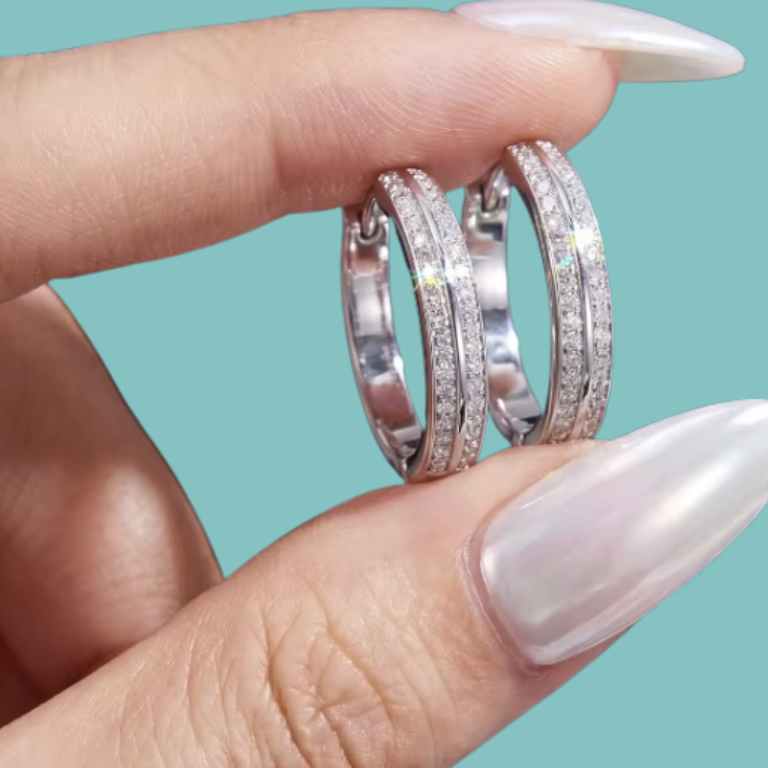 A close-up of a hand with long, glossy white manicured nails holding two silver hoop rings set with small clear stones against a solid light teal background.