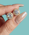 A close-up of a hand with long nude manicured nails holding a pair of round halo stud earrings. The earrings feature large clear center stones surrounded by smaller clear stones in a gold-tone setting, against a solid light aqua background.