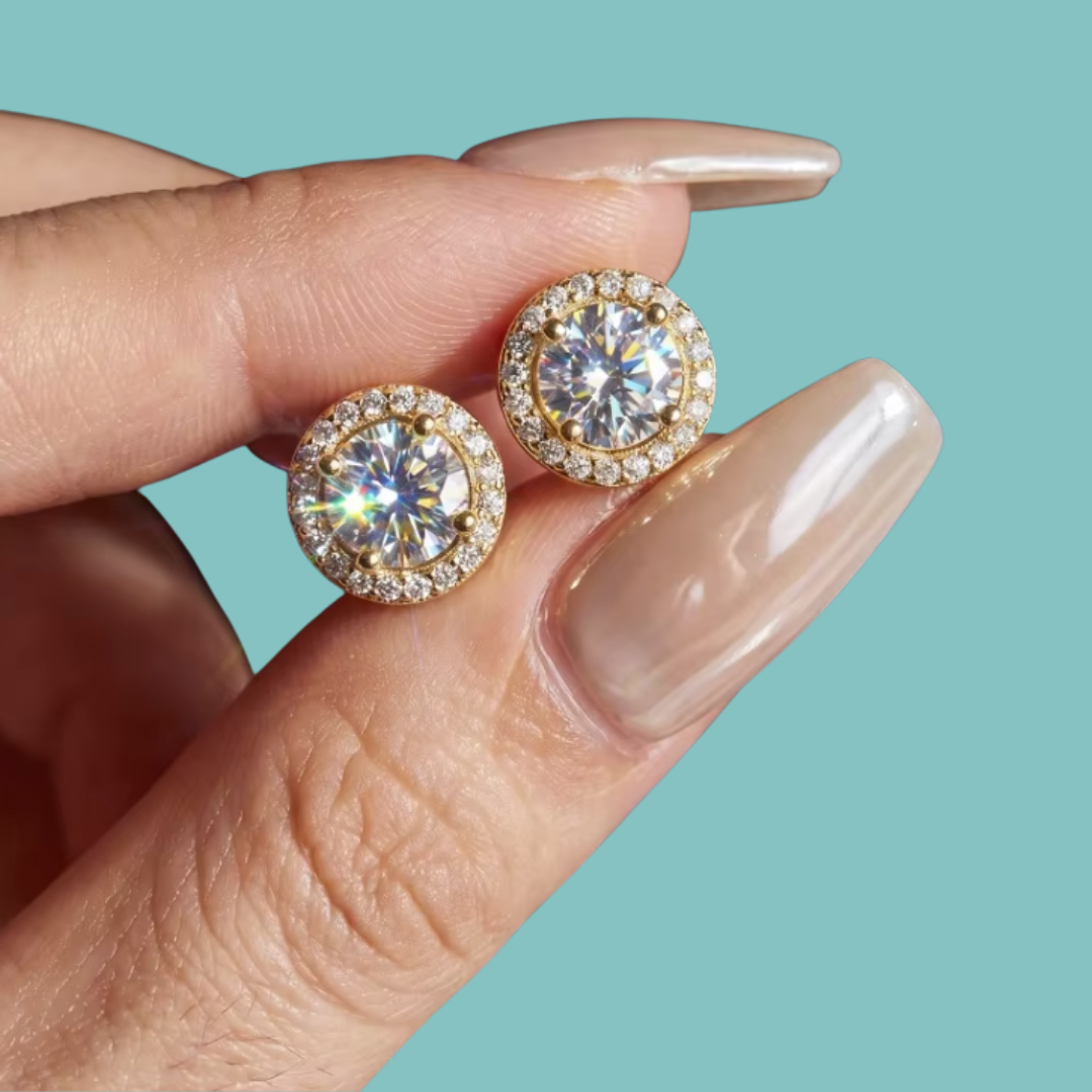 A close-up of a hand with long nude manicured nails holding a pair of round halo stud earrings. The earrings feature large clear center stones surrounded by smaller clear stones in a gold-tone setting, against a solid light aqua background.